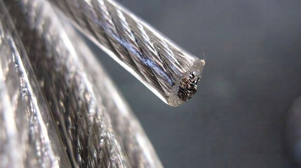 Como especificar o diâmetro do cabo de aço revestido 6mm para proteção e estaiamento? 2 Fotografia em macro de um cabo de aço revestido em corte, onde a ponta exposta revela os fios metálicos trançados da alma de aço envoltos por uma camada transparente e brilhante de PVC ou nylon. O foco está na textura dos fios e na espessura do revestimento. O fundo é um metal escuro desfocado. O cabo principal está enrolado em primeiro plano e desfocado.