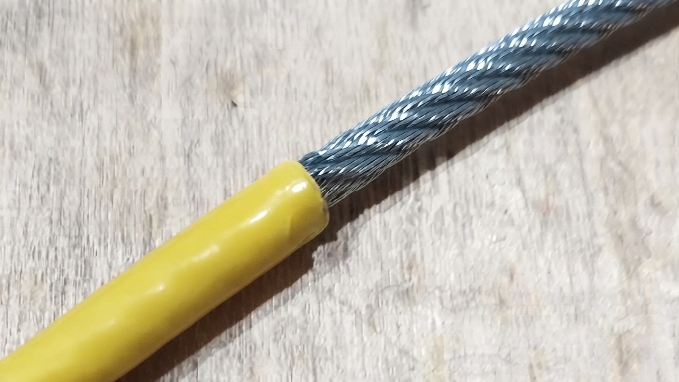 Detalhe de um cabo de aço revestido com capa amarela, mostrando a transição entre o cabo de aço galvanizado e o revestimento plástico sobre superfície de madeira.